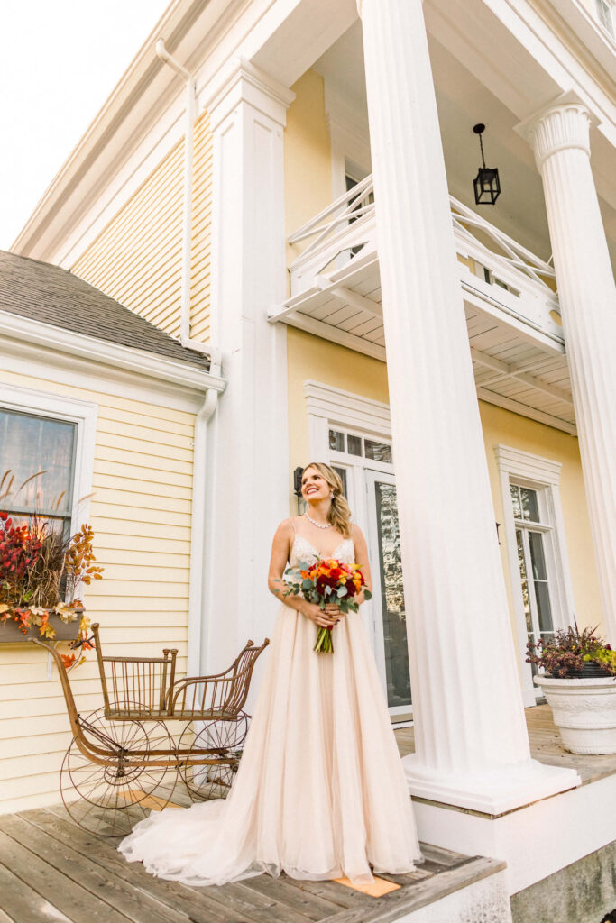 "A radiant bride in a beautiful flowing wedding dress holding a vibrant bouquet, standing on the porch of The Emerson House Inn in Orland, Maine, with a look of joyful anticipation, framed by classic white columns and the warm hues of an autumnal day."