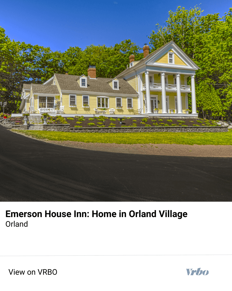 Alt text: "The Emerson House Inn, a picturesque yellow guest house with white trim and columns, nestled in Orland Village, Maine, under a clear blue sky. The image invites viewers to 'View on VRBO' for rental details."